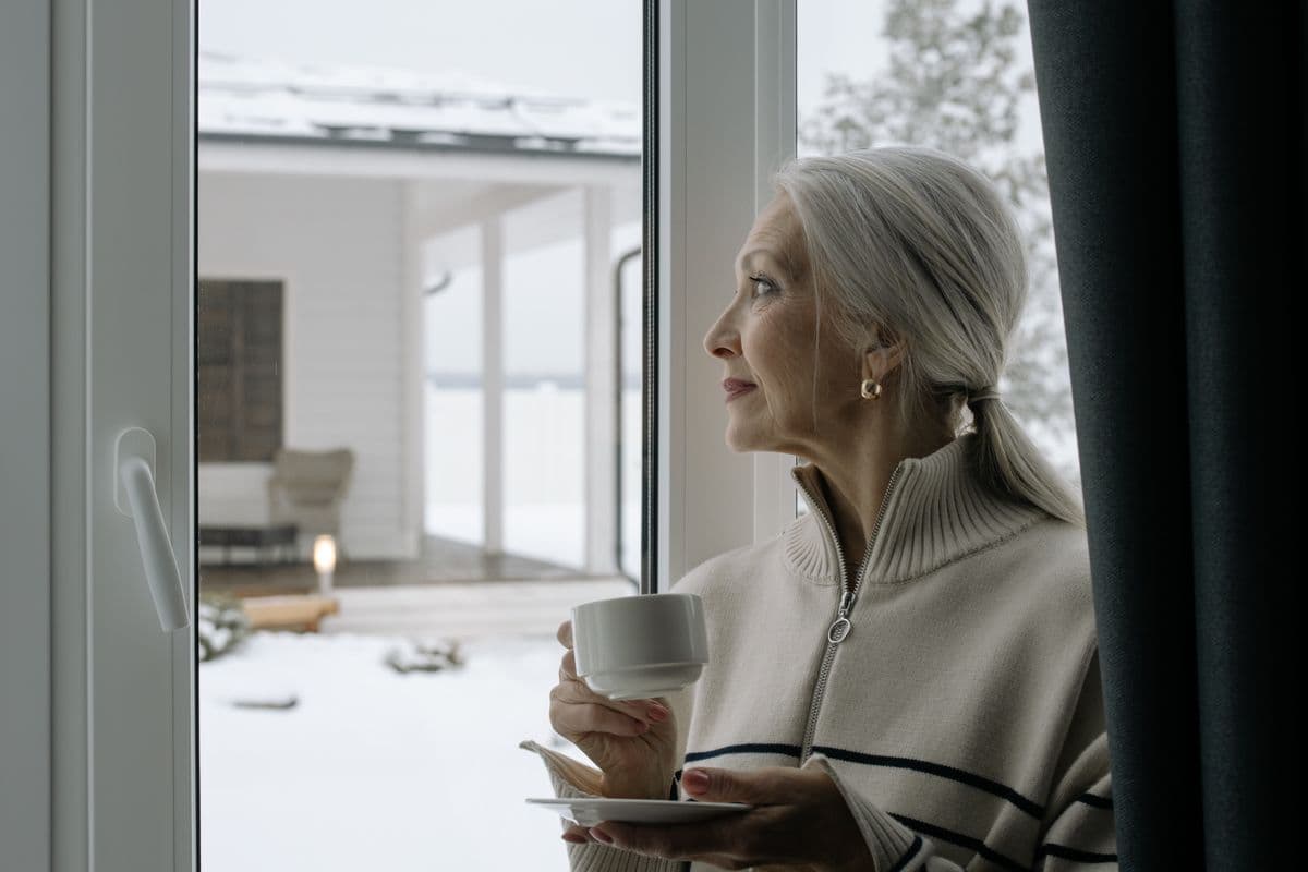 Elegant senior woman with gray hair holds a coffee cup, gazing out a window during winter.