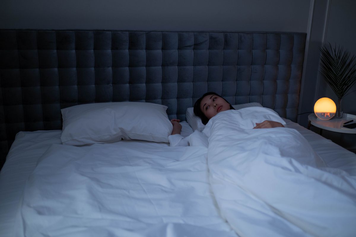 An Asian woman lying awake under soft white bedding in a dimly lit modern bedroom.
